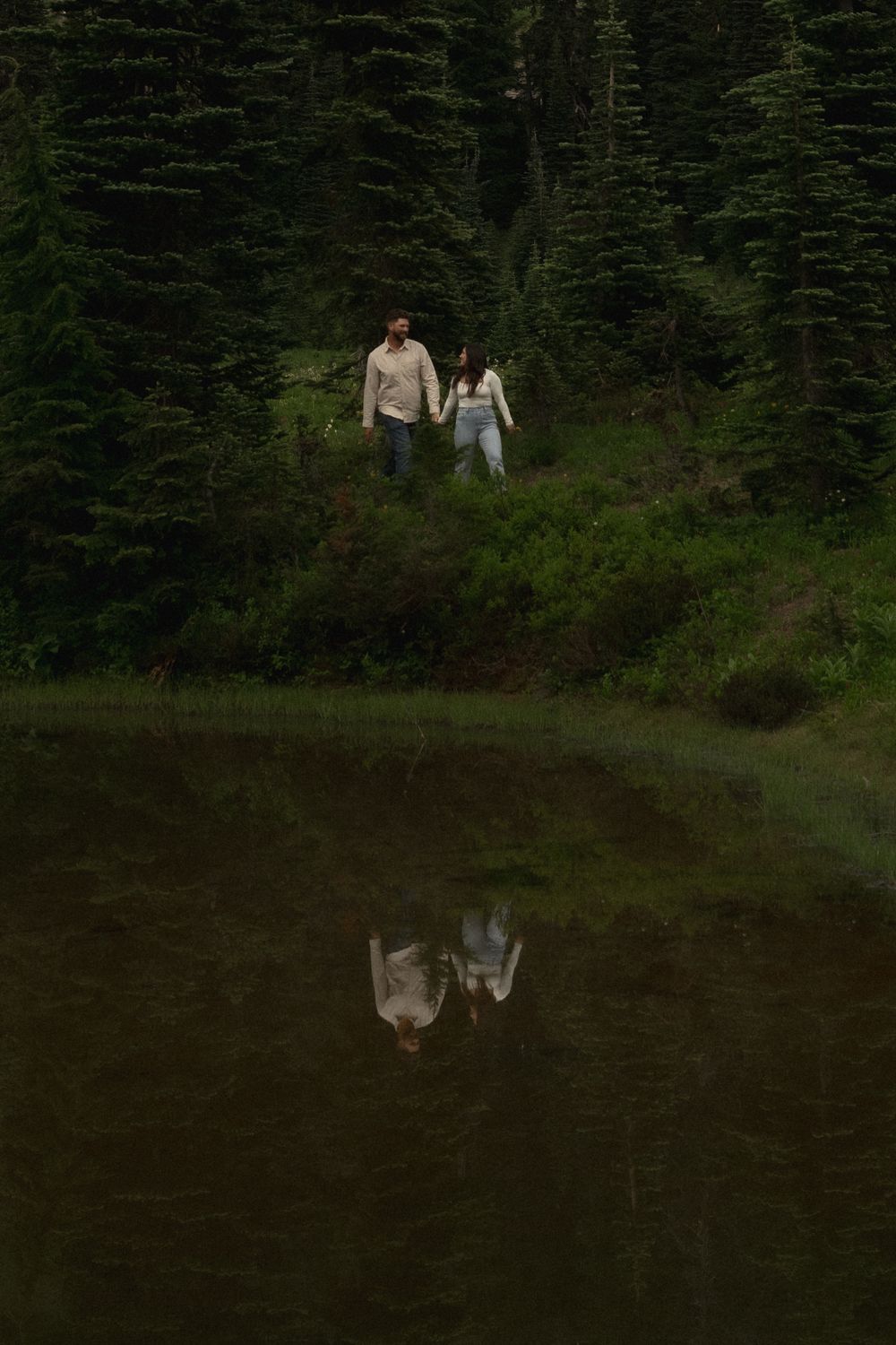Two people stand on a grassy slope near evergreen trees with their reflection mirrored in a dark, still lake below.