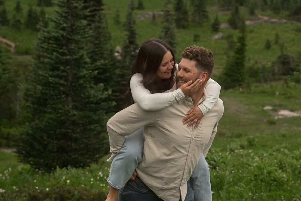 A couple embraces on a grassy mountainside surrounded by evergreen trees and wildflowers in a romantic moment.