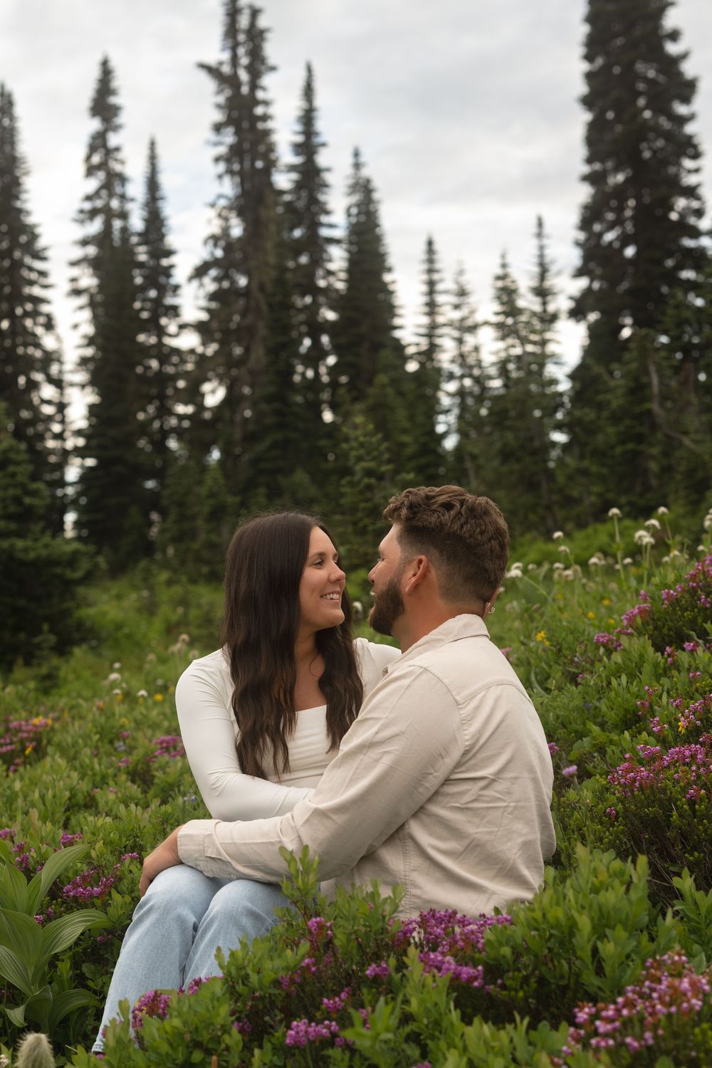Couple sitting together in a field of purple wildflowers with evergreen trees in the background during golden hour.