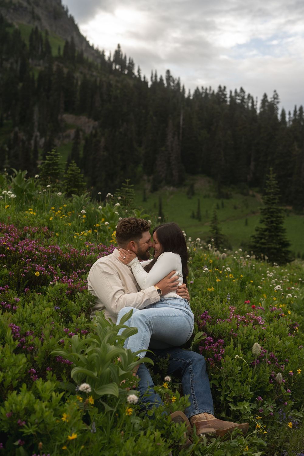 A couple embraces in a mountain meadow filled with colorful wildflowers under moody skies and evergreen trees.
