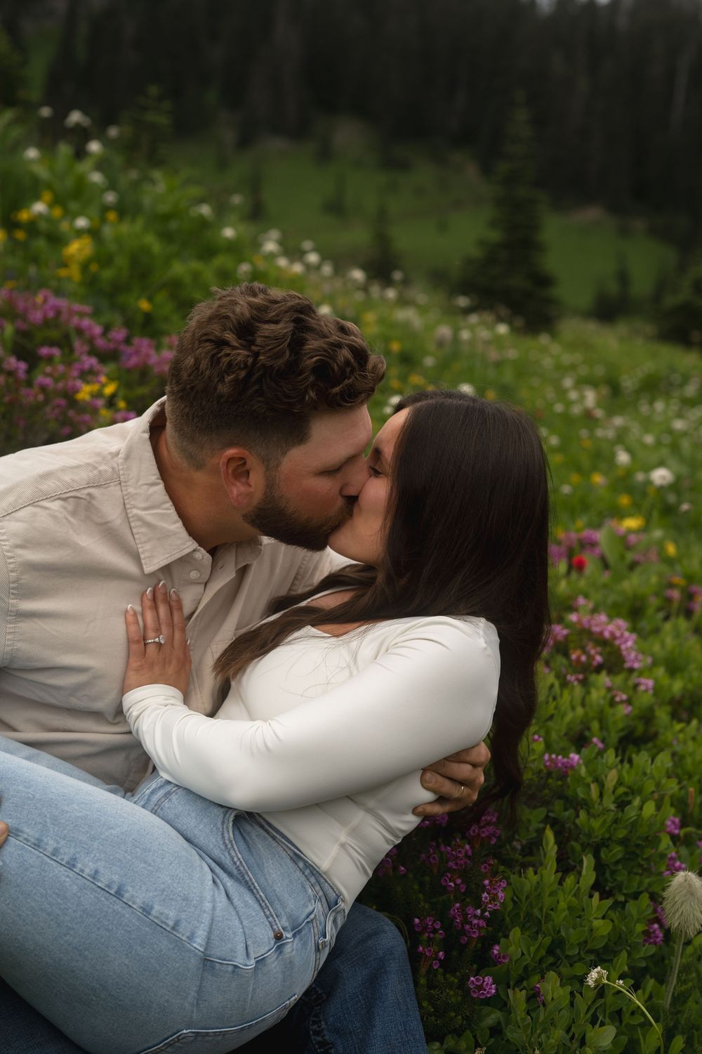 A romantic couple shares intimate kisses while sitting among colorful wildflowers in a mountain meadow.