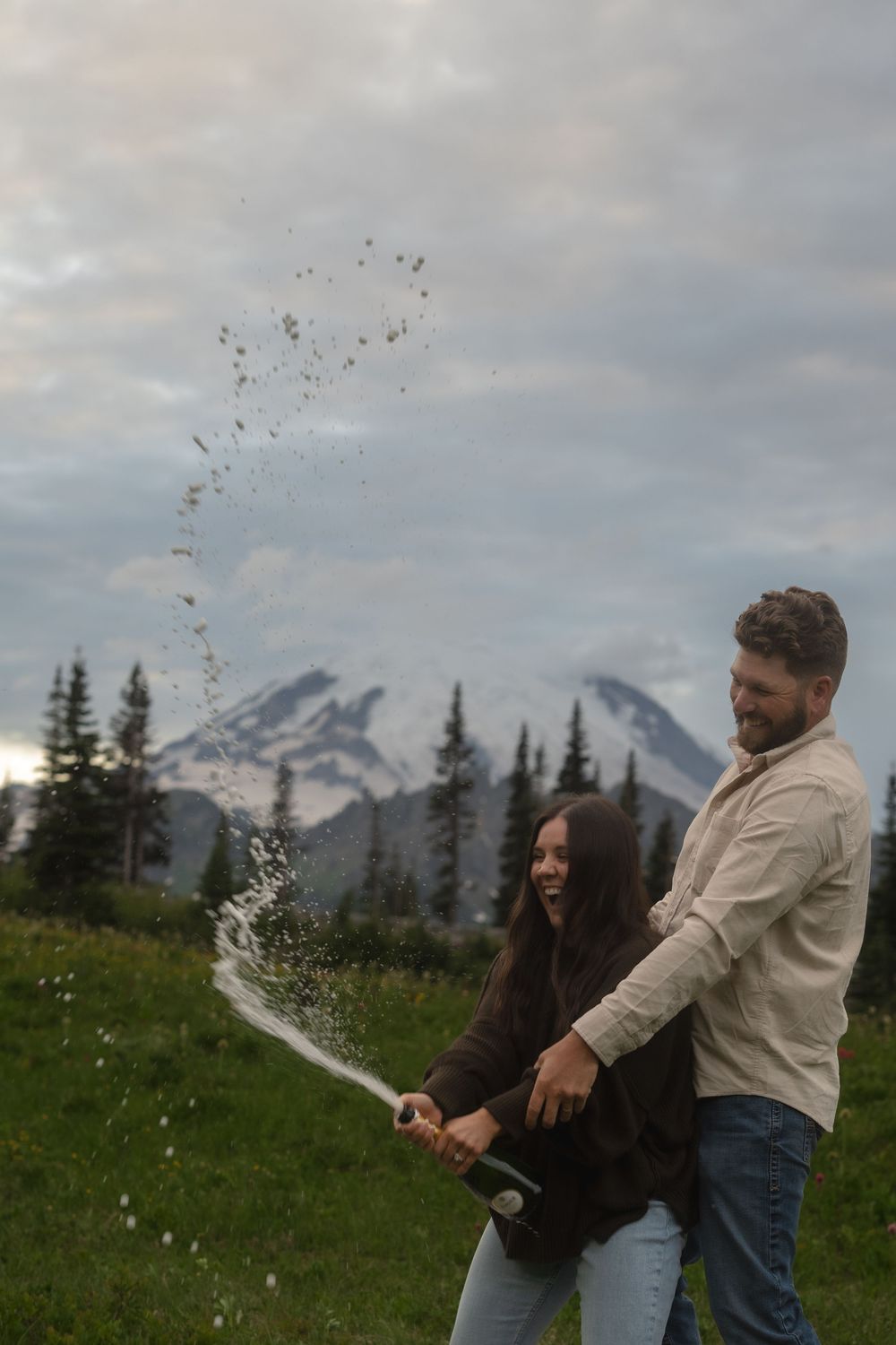 A couple pops champagne in a scenic mountain meadow with snow-capped peaks visible in the background.