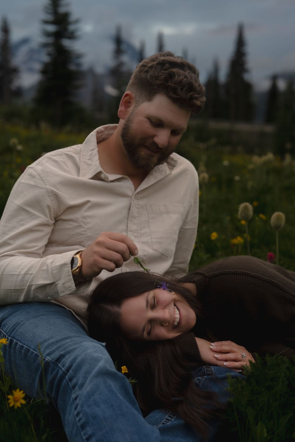 A couple sits together in a wildflower meadow at dusk with evergreen trees in the background.