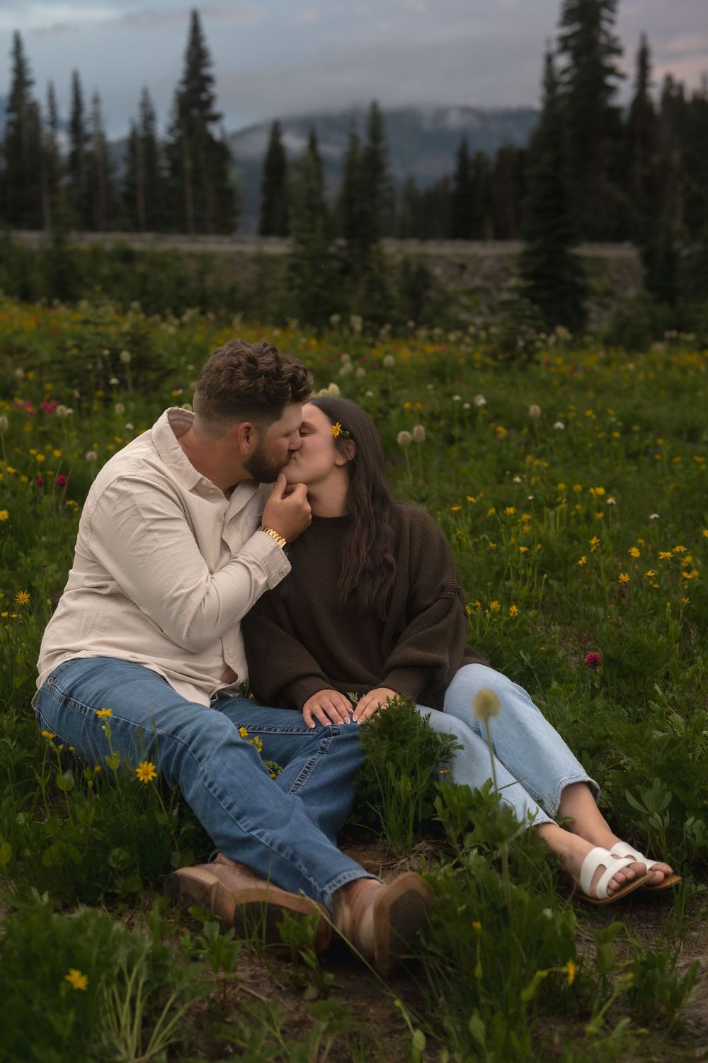 Couple sitting together in a mountain meadow filled with yellow wildflowers and evergreen trees at dusk.