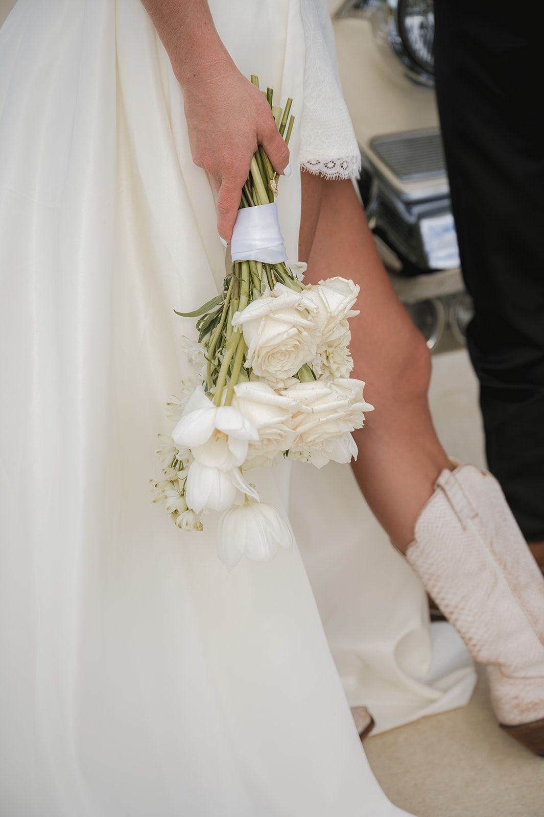 A bride wearing a white dress holds a delicate bouquet of white flowers with greenery while wearing western boots.