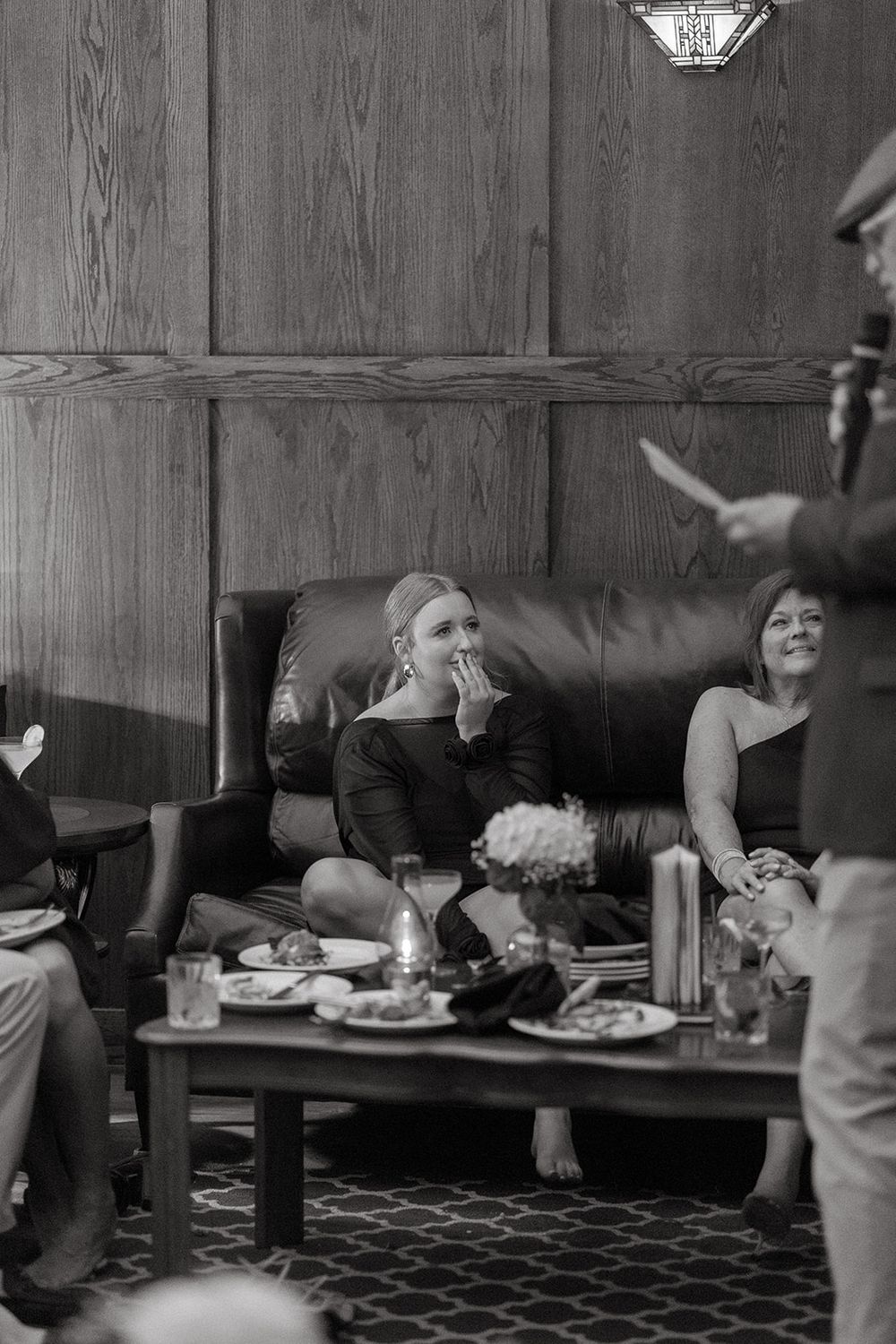 Black and white photo of guests relaxing on a leather couch at a social gathering with food and drinks on the table.