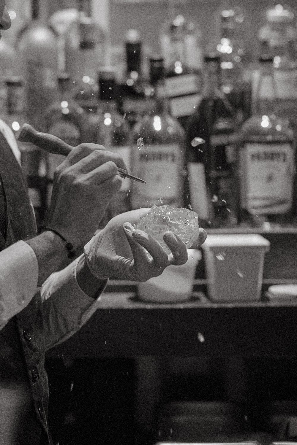 Bartender's hands mixing cocktails at a dimly lit bar counter with bottles visible in the background.