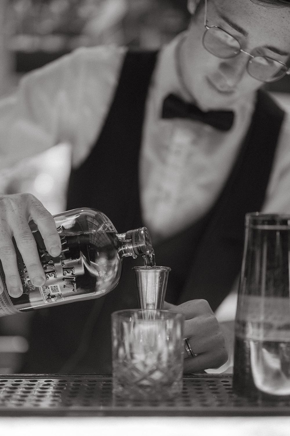A black and white image shows hands pouring drinks at a bar with crystal glasses and cocktail equipment.