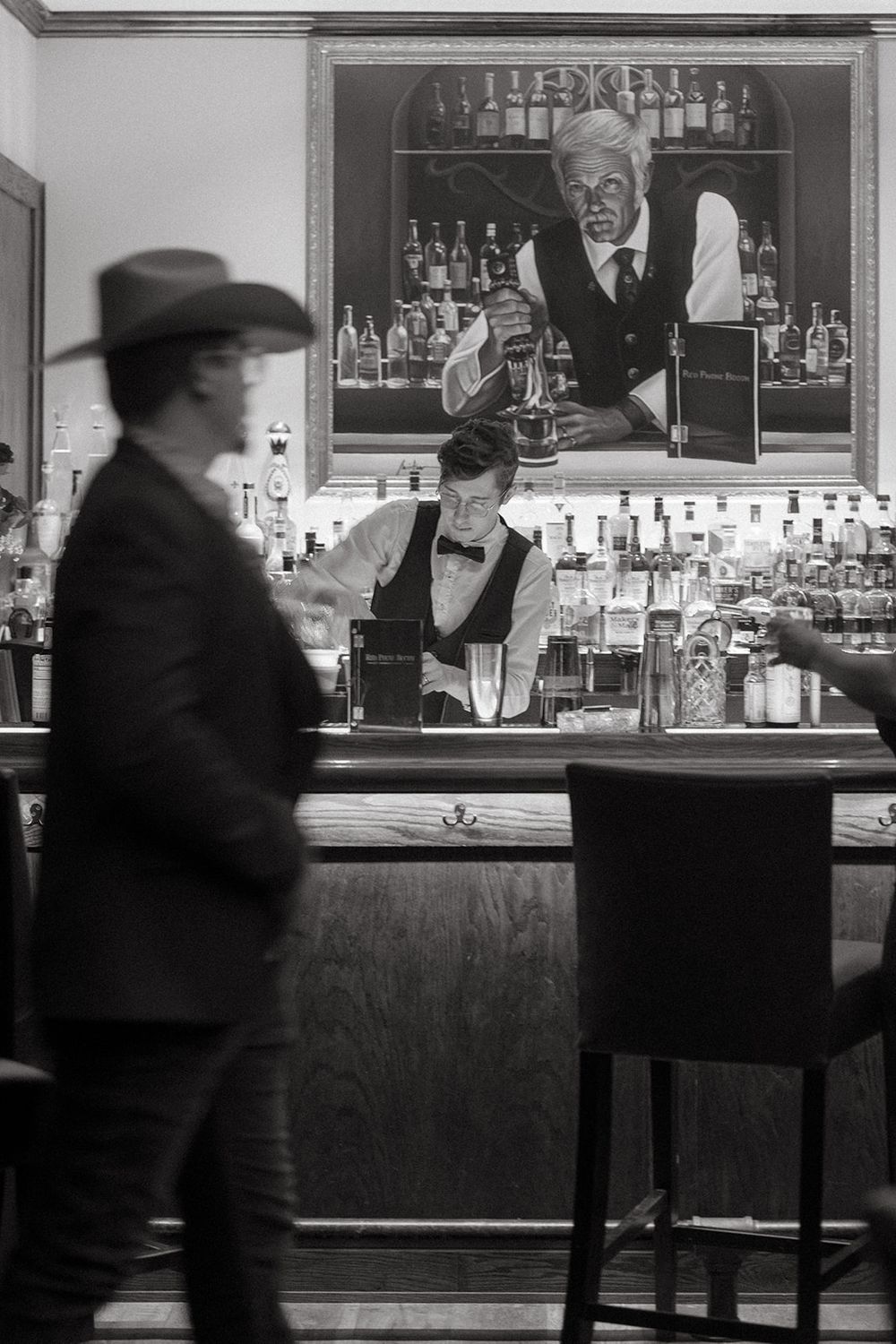 A black and white panoramic shot of a vintage bar interior with patrons sitting at stools along the counter.
