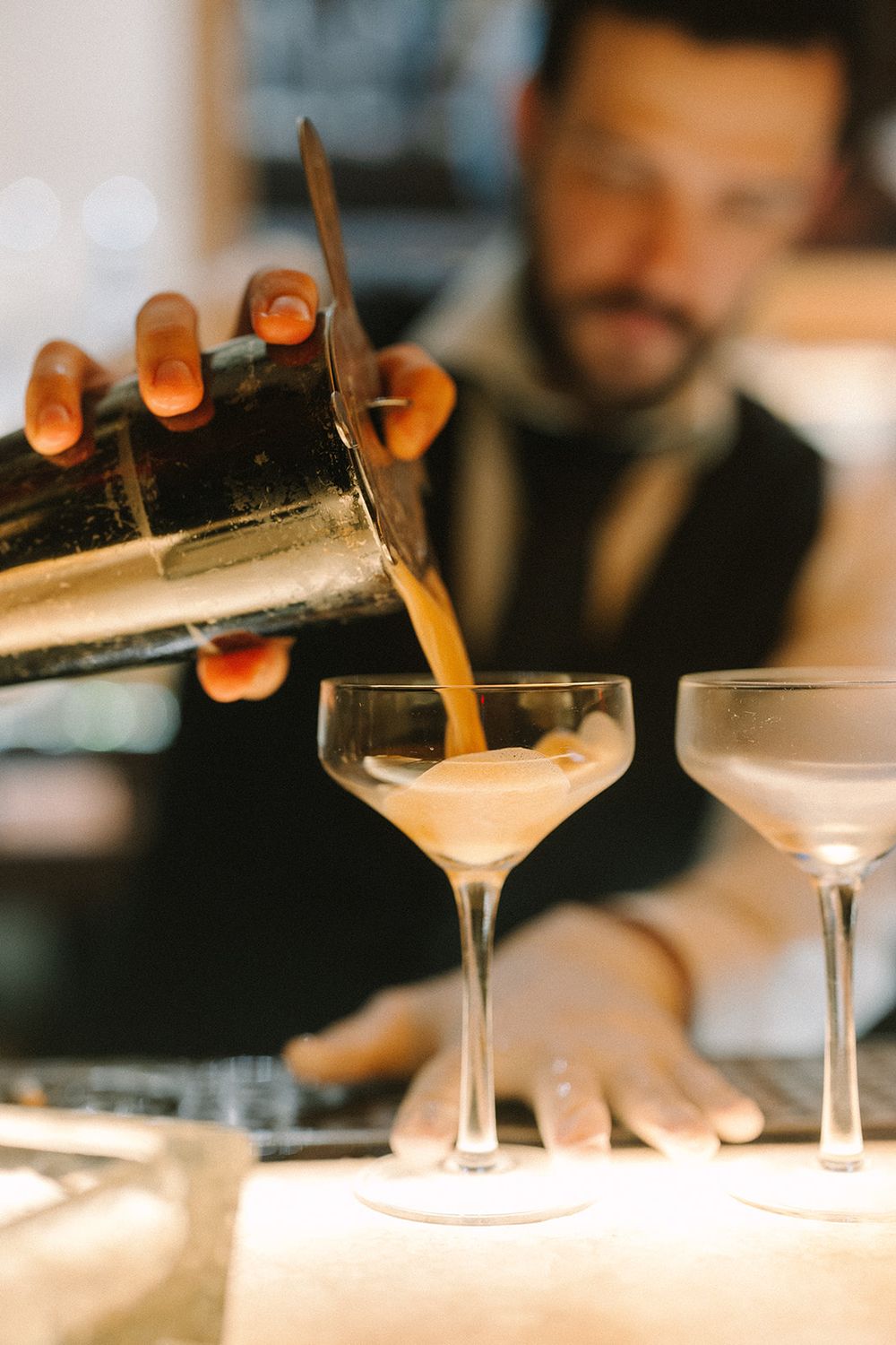 A bartender pours a creamy cocktail from a shaker into elegant coupe glasses at an upscale bar.