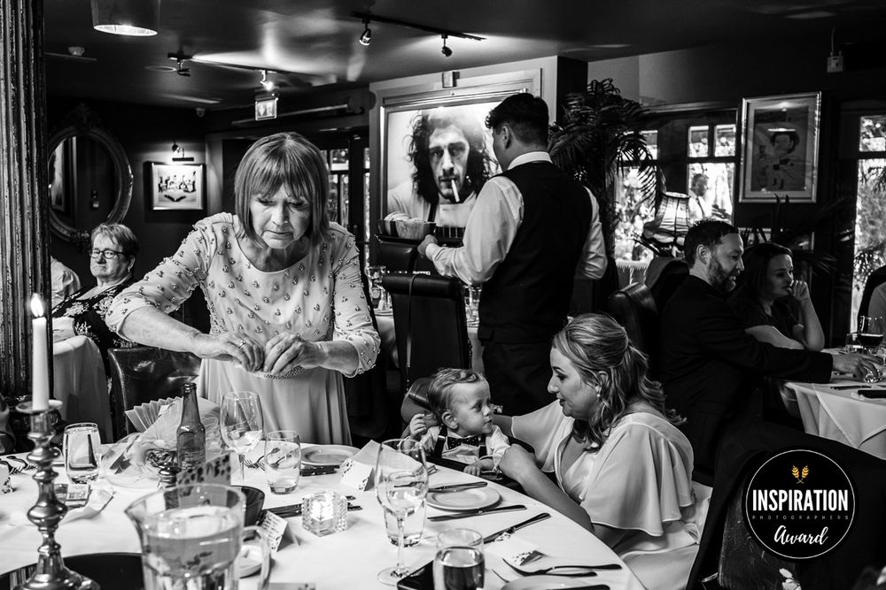 A black and white candid restaurant scene shows diners and staff interacting at a busy table with drinks and dishes.