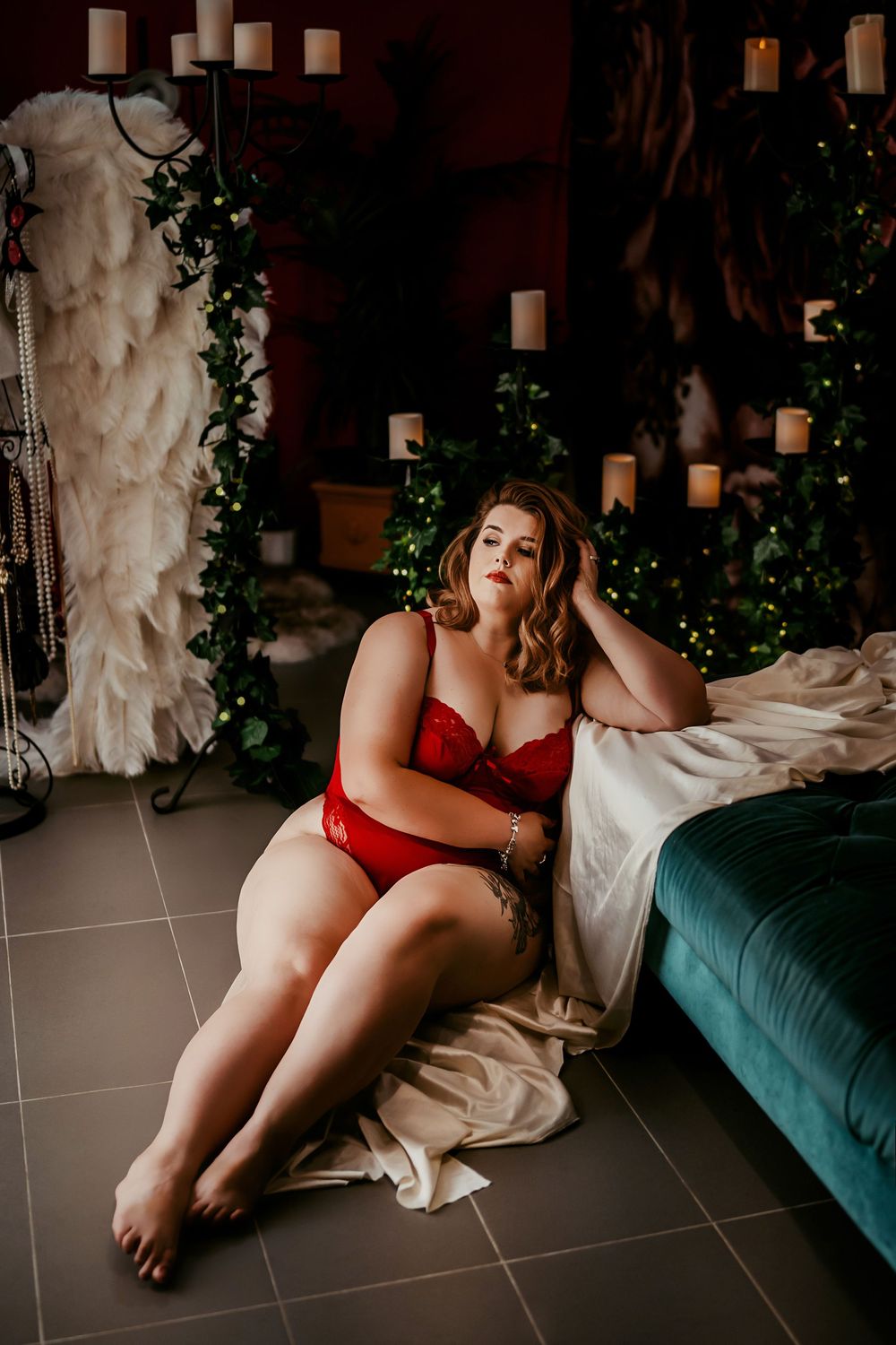 Cozy holiday boudoir setting with red lingerie, white candles, greenery and velvet furniture against dark background.