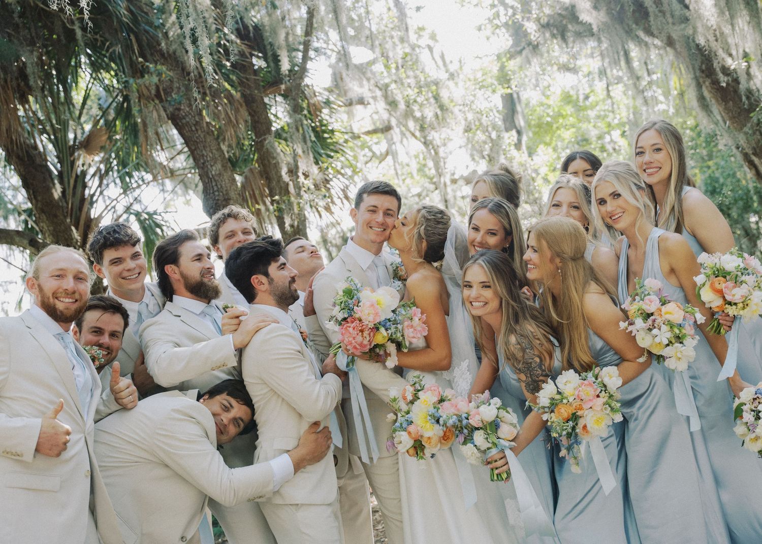 A joyful wedding party celebrates under Spanish moss-draped trees with bridesmaids in blue gowns.
