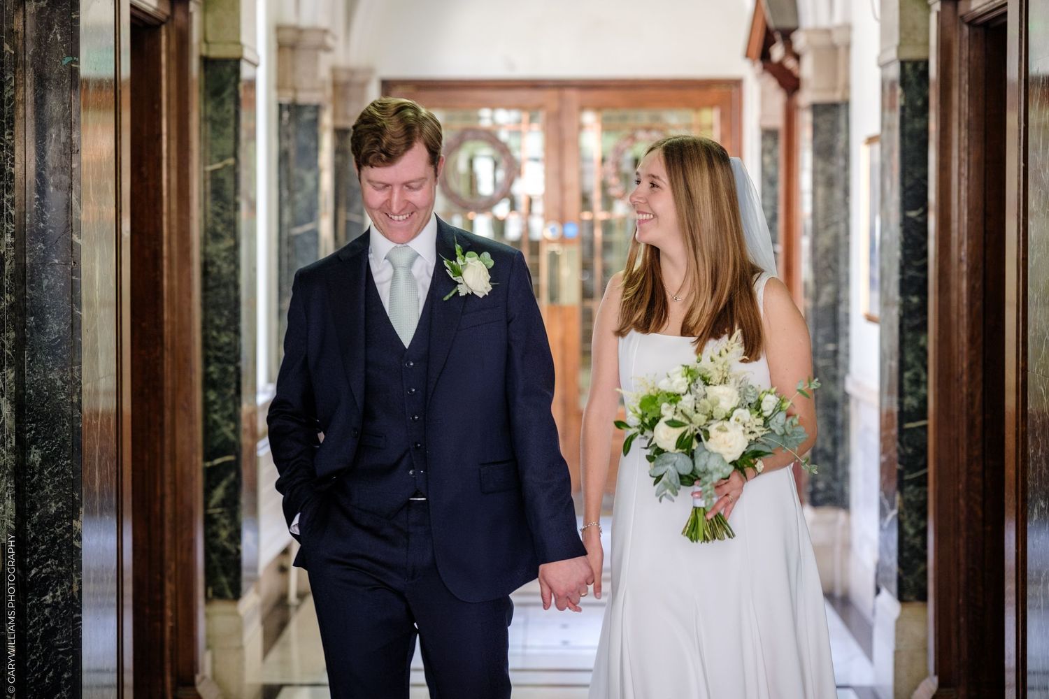 A couple walks down an elegant corridor holding hands on their wedding day, with the bride carrying a white bouquet.