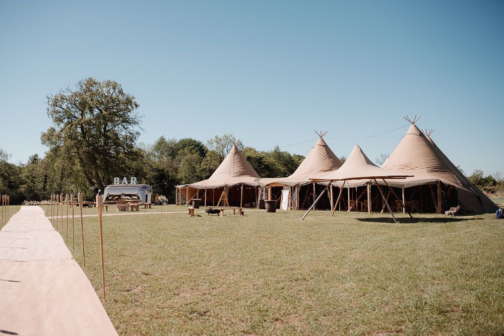 Three large brown tipi-style tents set up on green grass field with trees in background under blue sky.