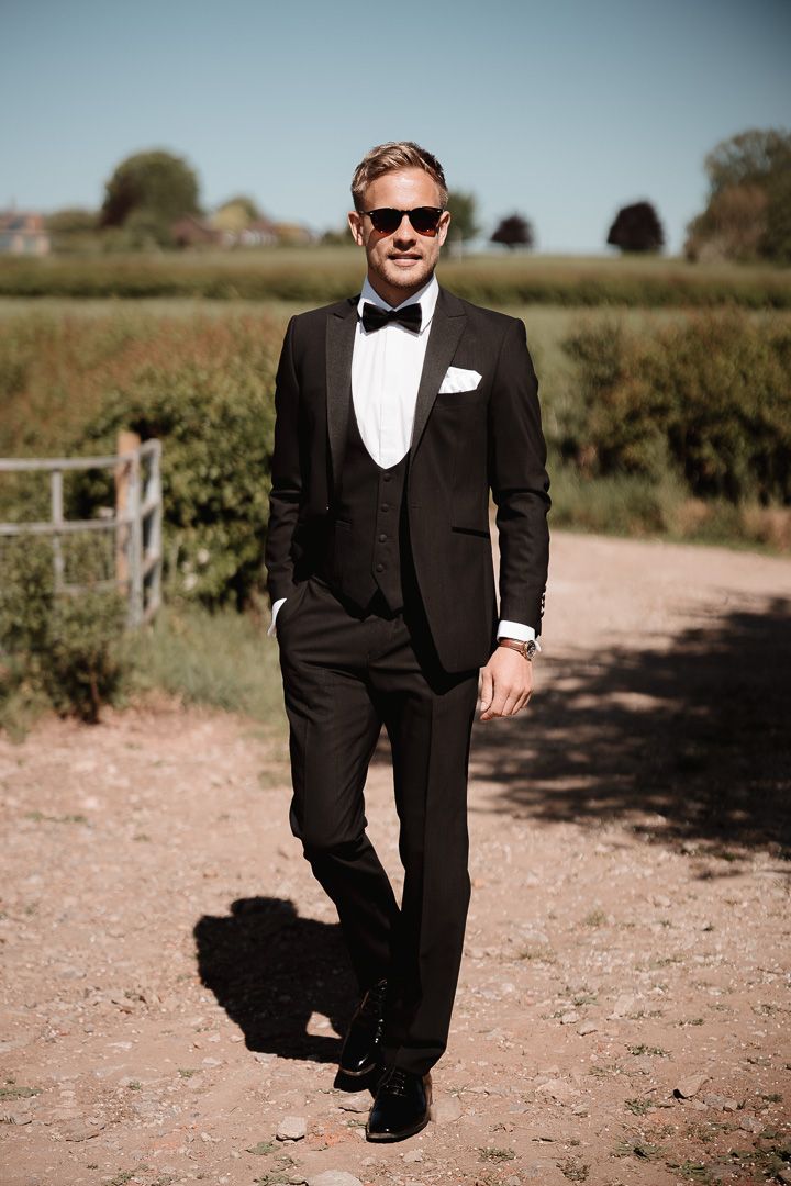 A person in a classic black tuxedo with bow tie and sunglasses poses on a dirt path in the countryside.