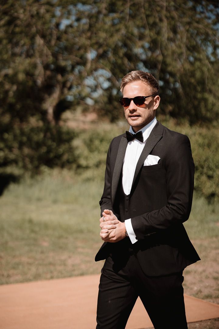 A groom in a classic black tuxedo, bow tie and sunglasses poses outdoors on a sunny day against a natural backdrop.