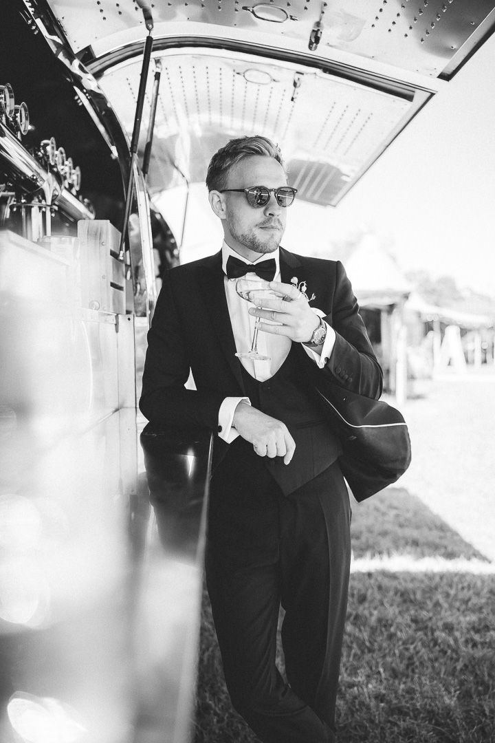 Elegant black and white photo showing figure in tuxedo and bow tie holding drink on vintage train car.