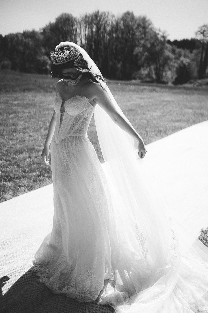 A bride in a flowing white dress and veil stands in a sunlit field with trees in the background, captured in black and white.