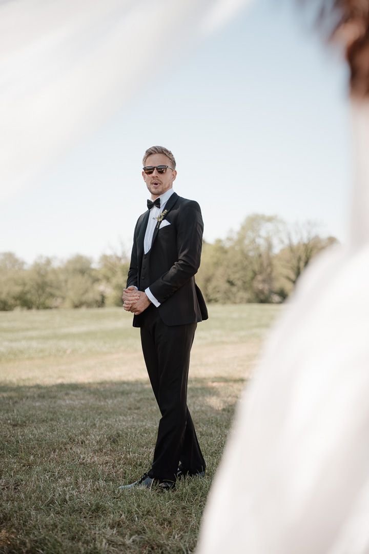 A groom in a classic black tuxedo and sunglasses stands in a grassy field during an outdoor wedding ceremony.