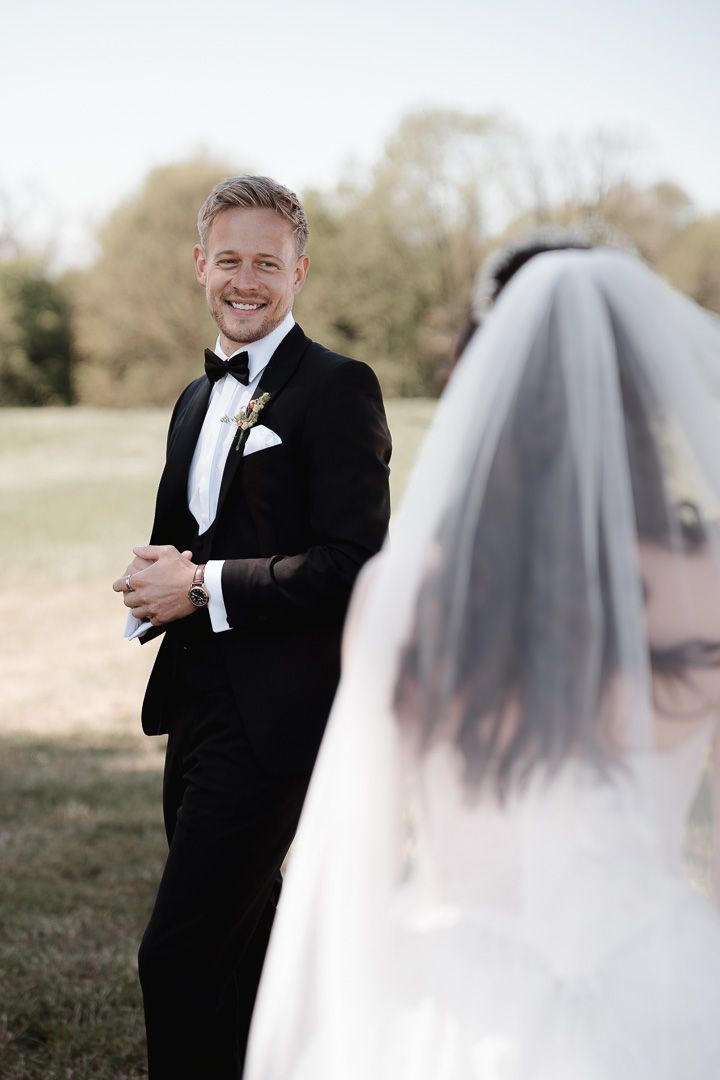 A groom in a classic black tuxedo and bowtie smiles while standing outdoors on his wedding day during a first look.