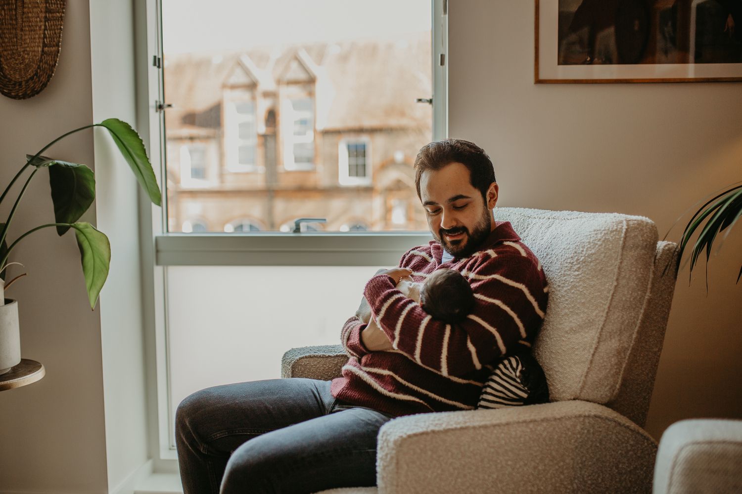 Person in a cozy armchair laughs while cuddling a newborn baby.