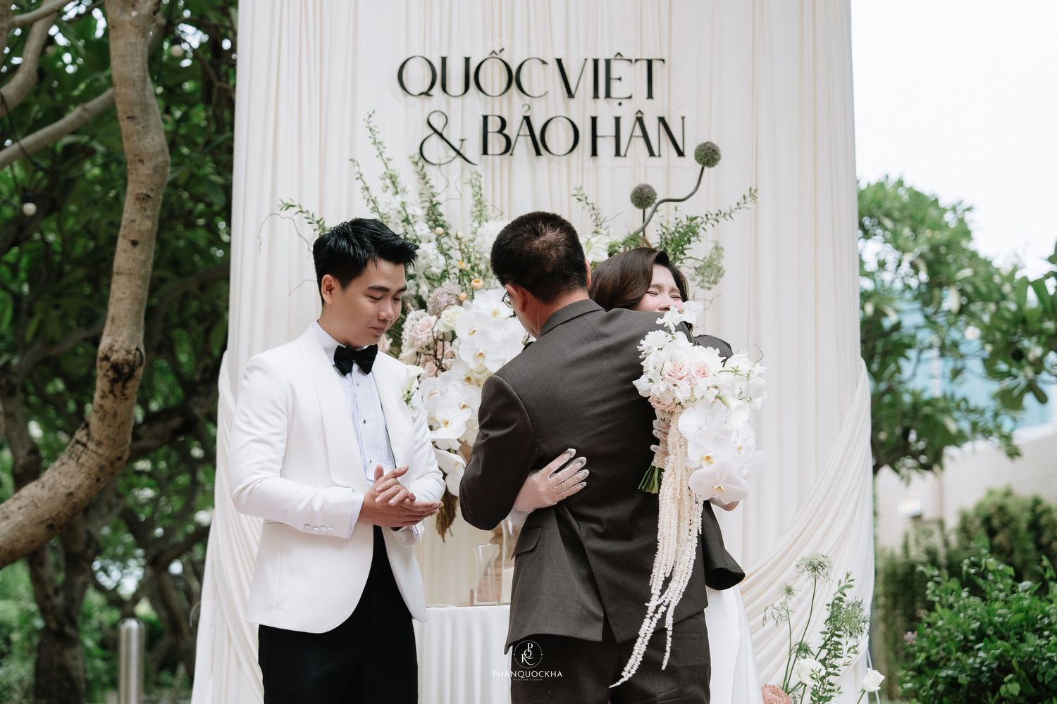 Groom in white tuxedo greets guest at elegant outdoor wedding ceremony with floral backdrop.