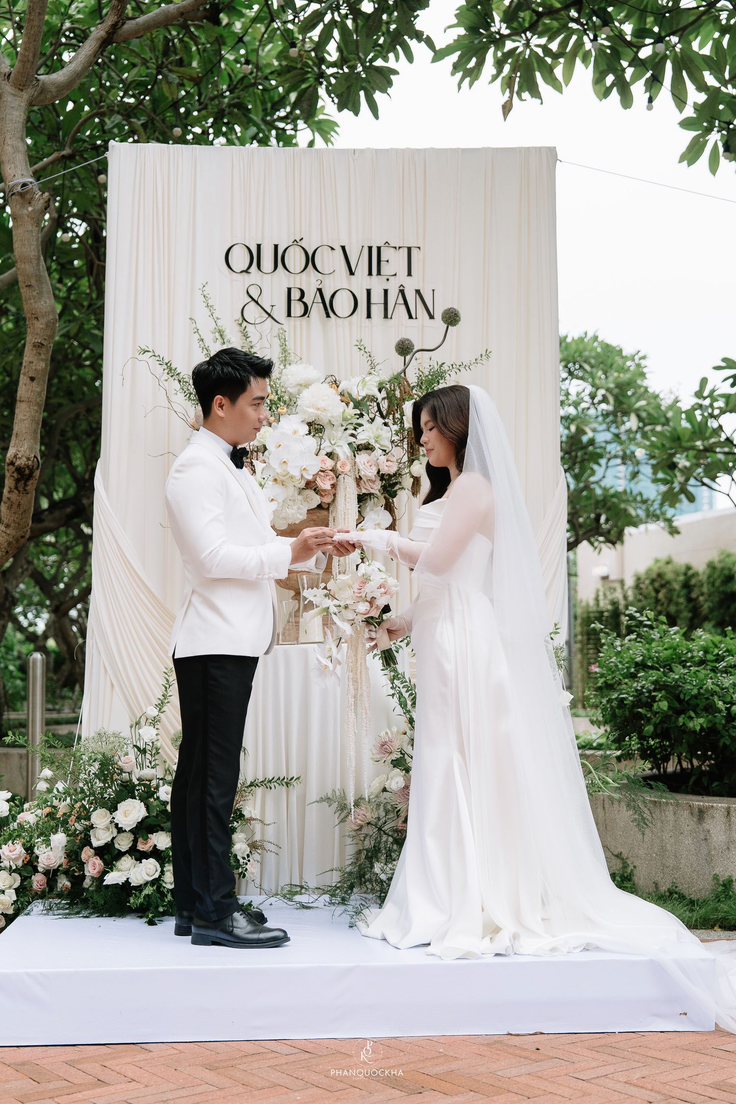 Couple exchanges rings during outdoor wedding ceremony before elegant floral backdrop.