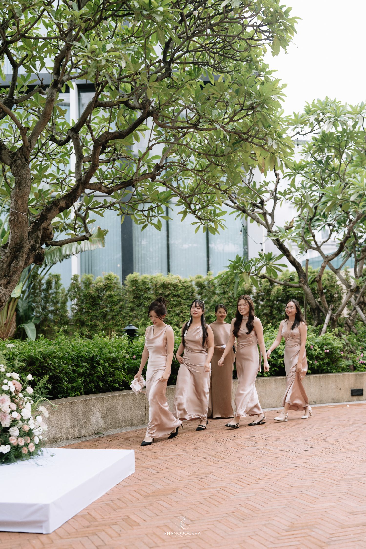 Five bridesmaids in champagne satin dresses walking together through a lush outdoor garden wedding venue.