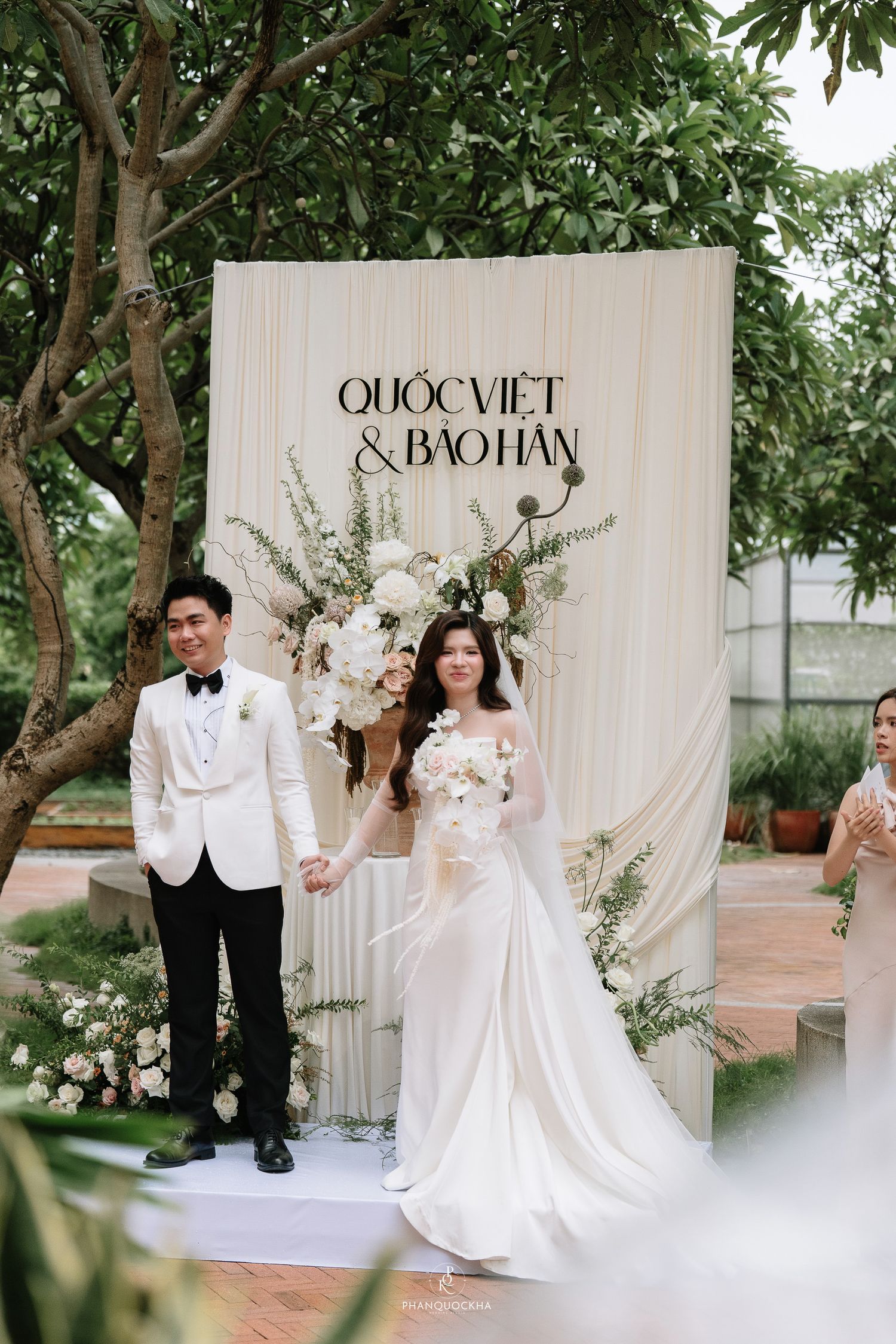 Newlyweds Quoc Viet and Bao Han hold hands smiling before elegant white floral wedding backdrop outdoors.