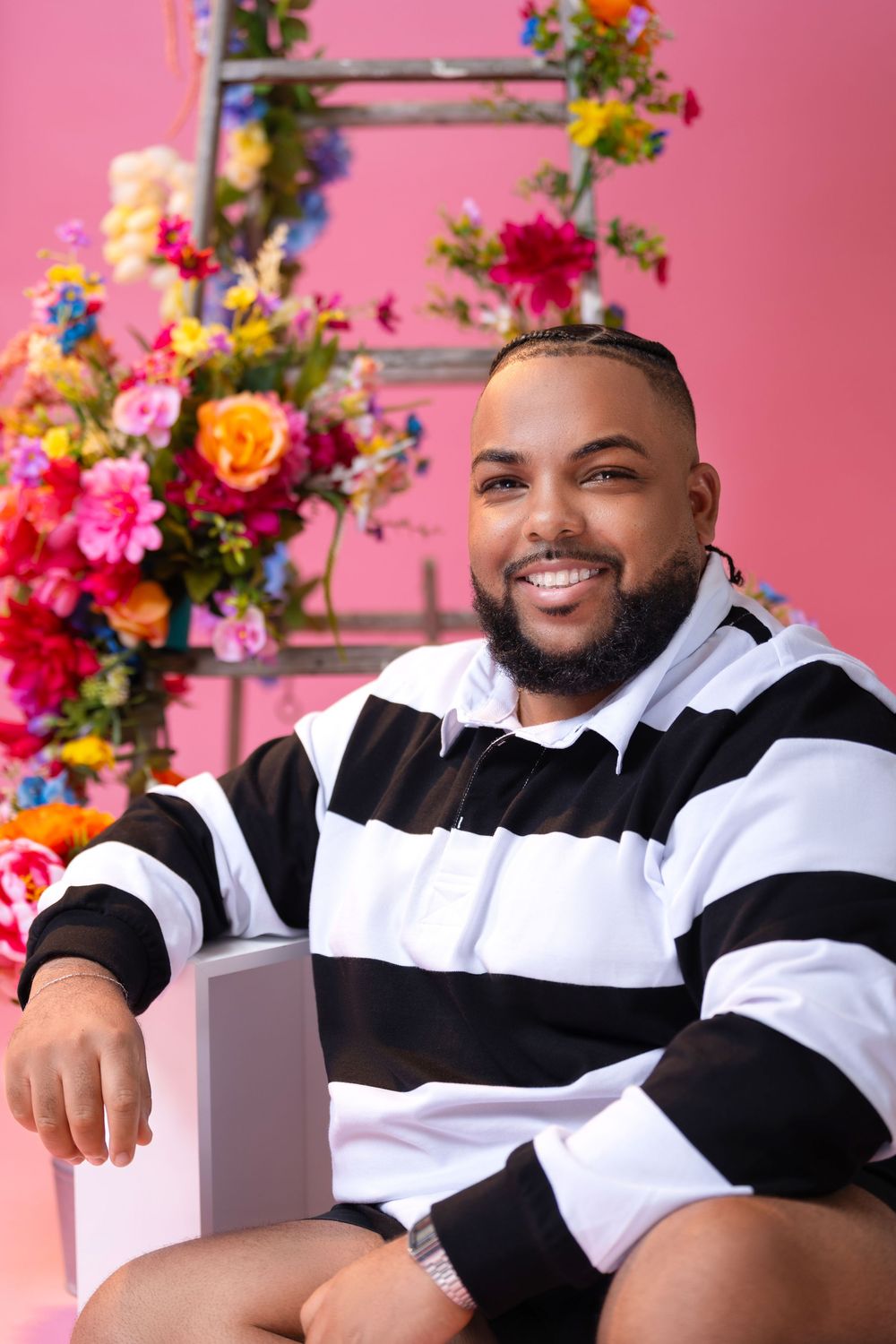 A smiling figure poses confidently in black and white athleisure wear against a vibrant pink backdrop with colorful flowers.