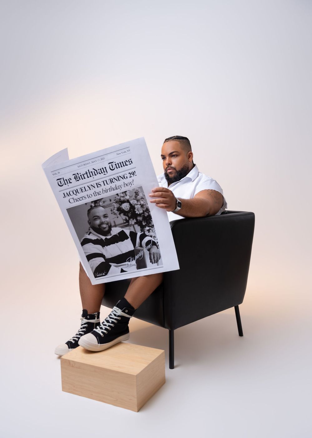 A person sits in a black armchair reading a newspaper with a wooden footrest in a minimalist white setting.