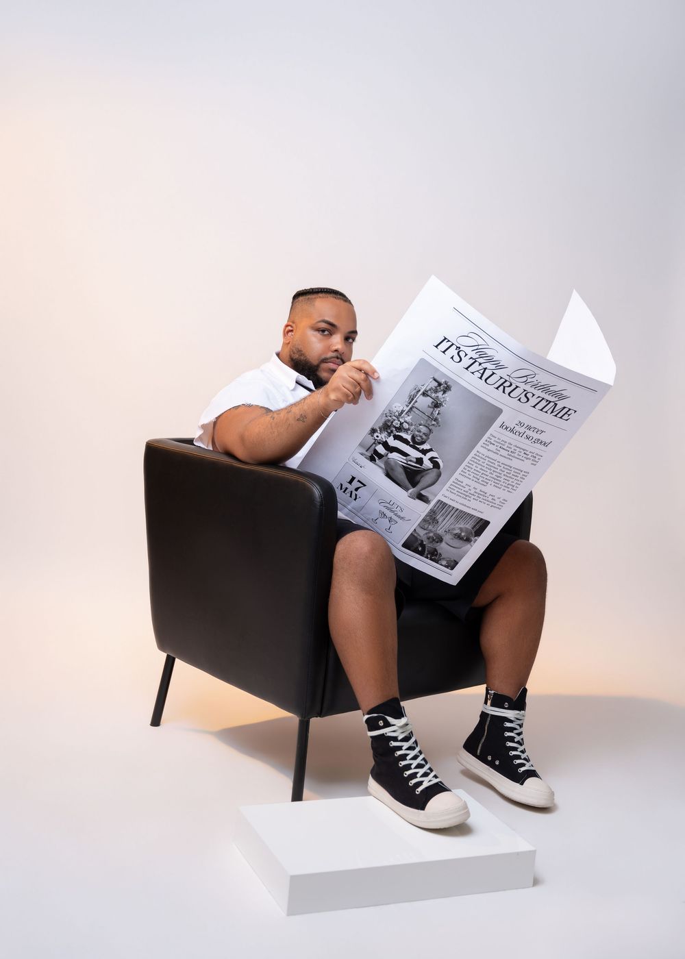A reader sits thoughtfully in a black armchair while reading a newspaper, wearing high-top sneakers and resting feet on white pedestal.