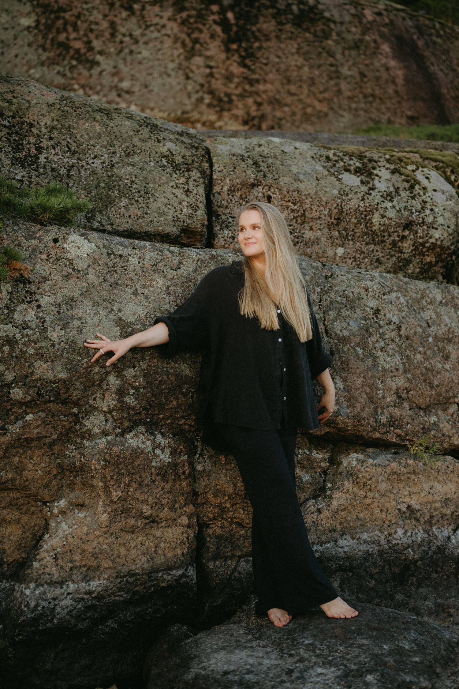 Figure in flowing black dress poses dramatically against large weathered rock formation at dusk.