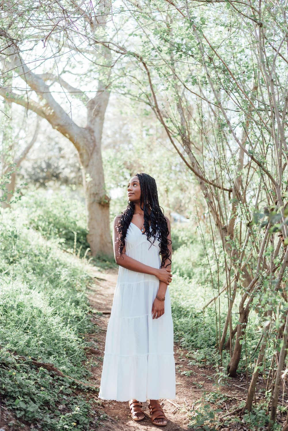Woman in flowing white maxi dress stands on nature trail surrounded by lush green trees in ethereal forest setting.