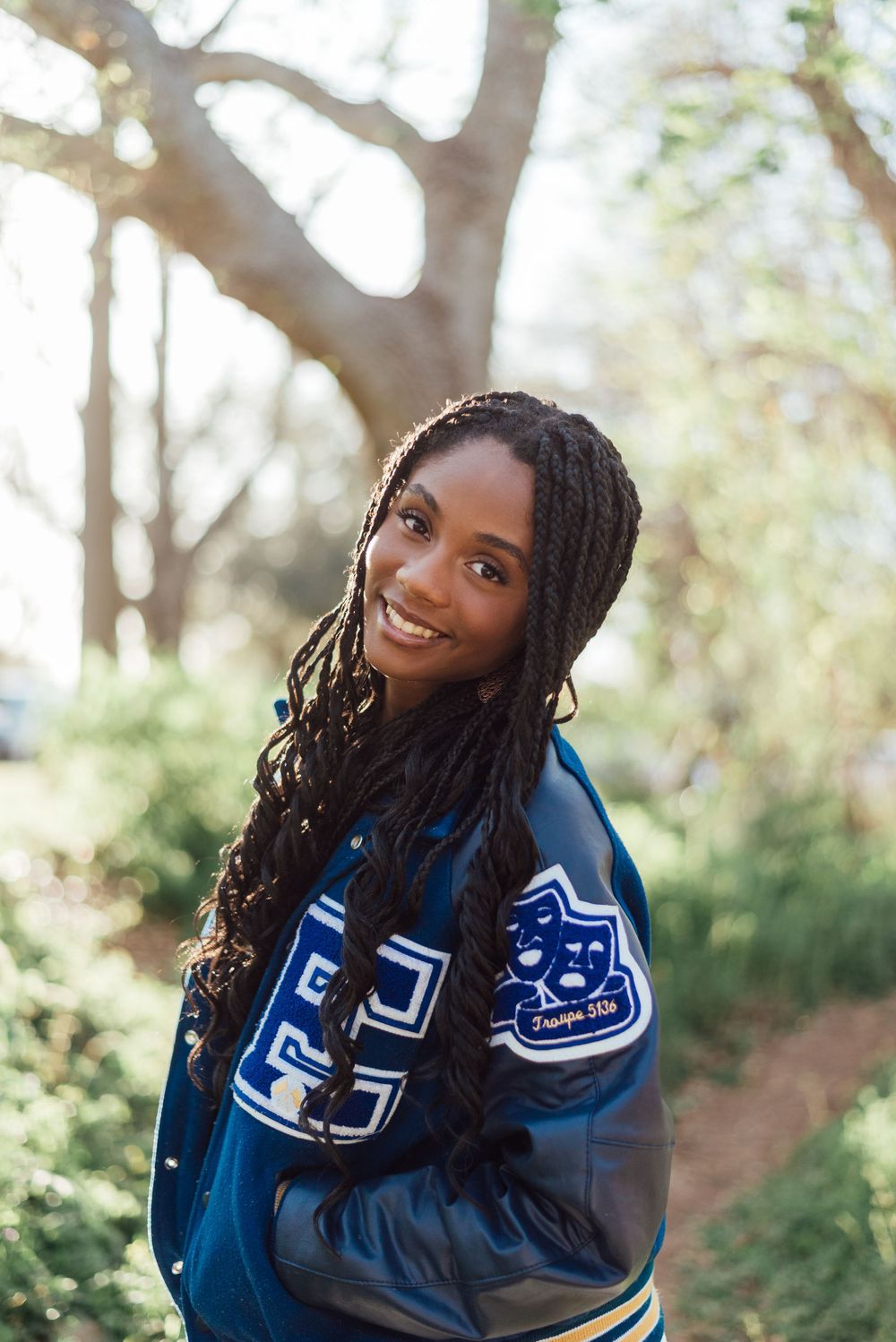Person wearing a blue varsity letterman jacket smiles outdoors in a sunlit wooded setting.