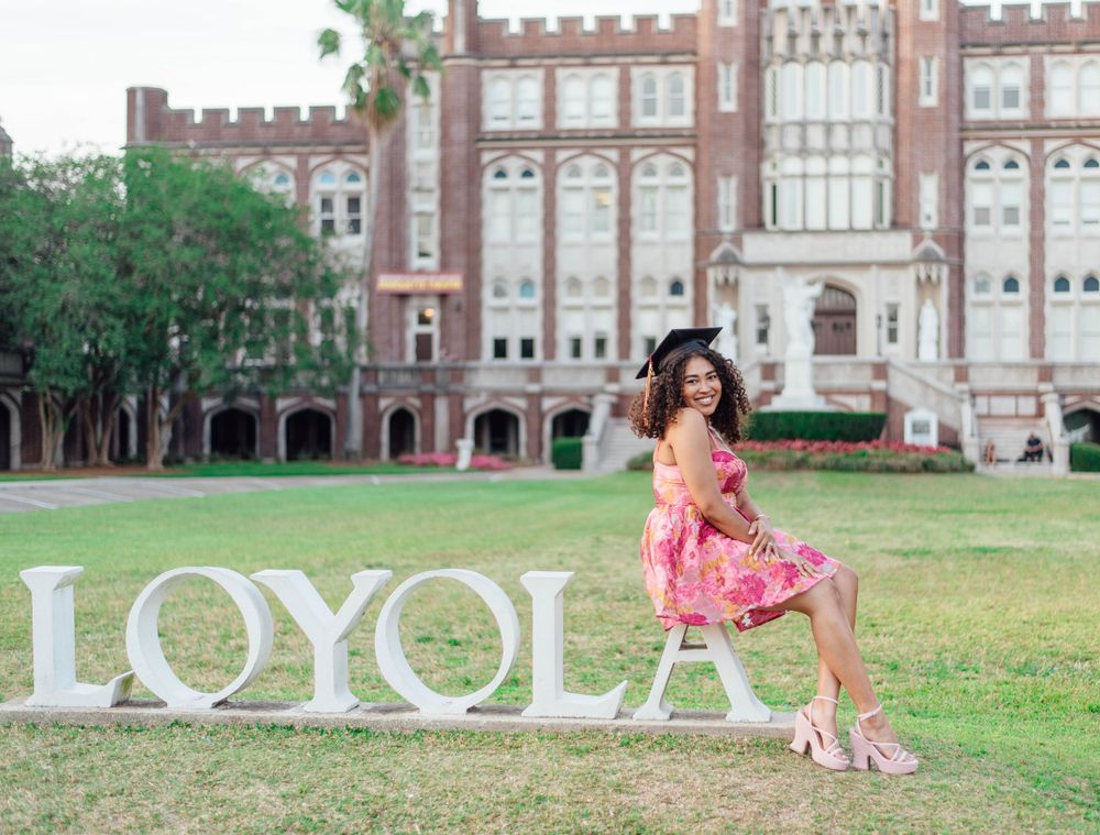 A person in a pink floral dress poses sitting on large white LOYOLA letters in front of a brick collegiate building.