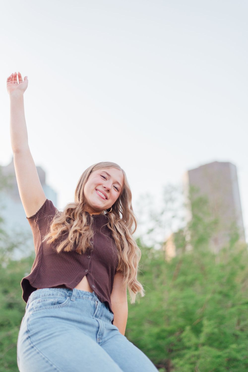 Person in a brown top and light jeans poses joyfully outdoors with arm raised against a blurred cityscape background.
