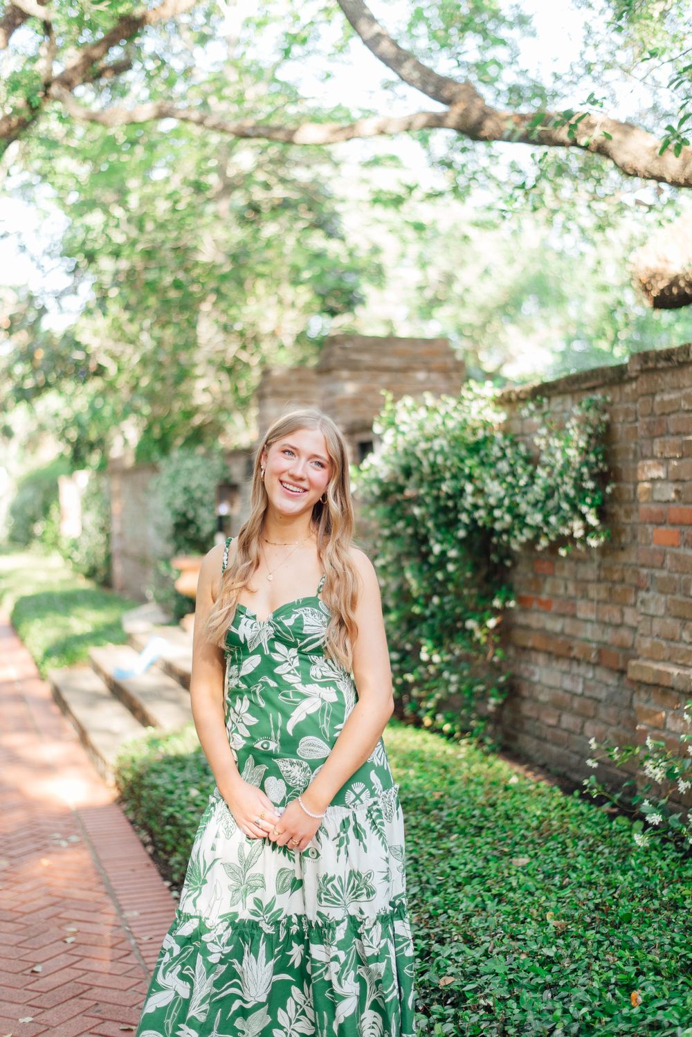 A person wearing glasses and a green and white tropical print dress poses in a lush garden setting with brick walls.