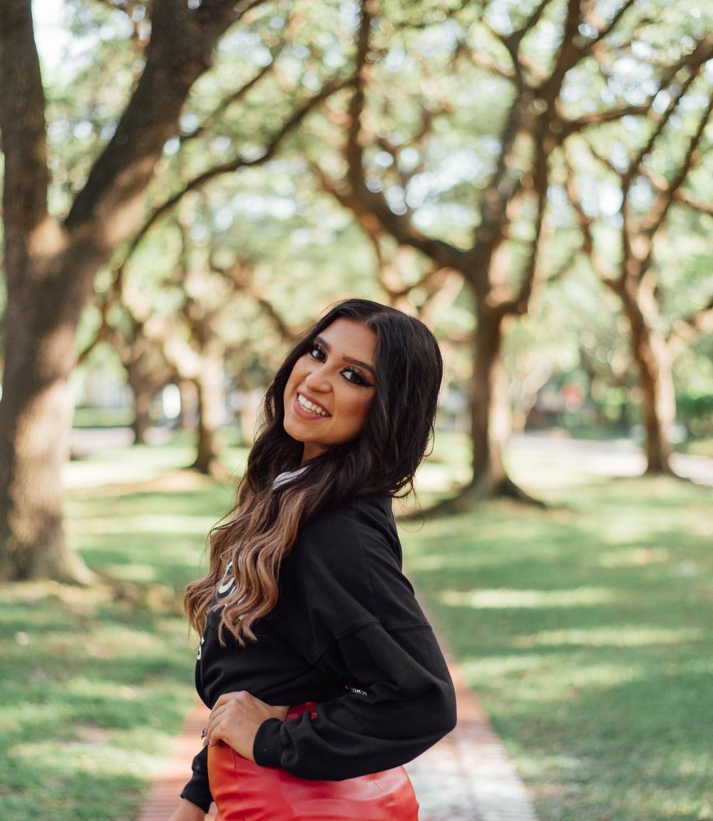 Woman in a black sweater and red skirt smiling while standing outdoors in a park with sunlit trees in the background.
