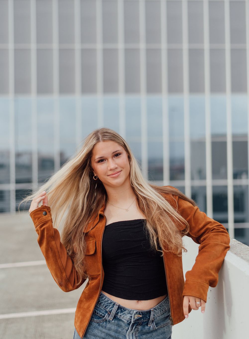Young woman in brown suede jacket and black crop top poses on rooftop parking structure on cloudy day.