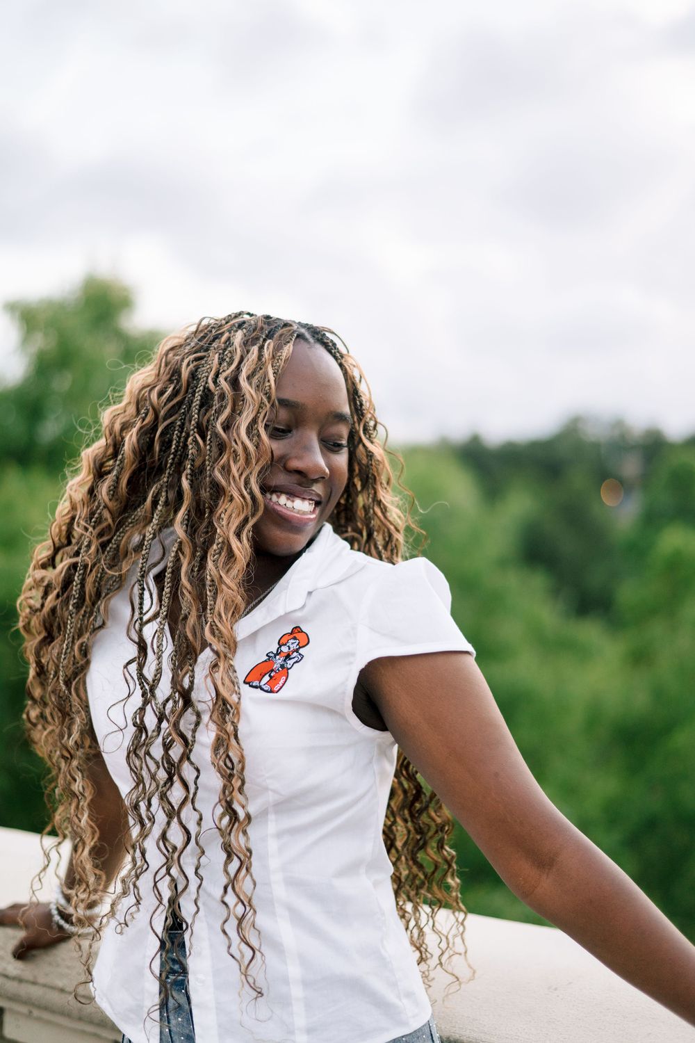 Young Black woman with long curly highlighted hair smiles outdoors, wearing a white polo shirt with colorful logo.