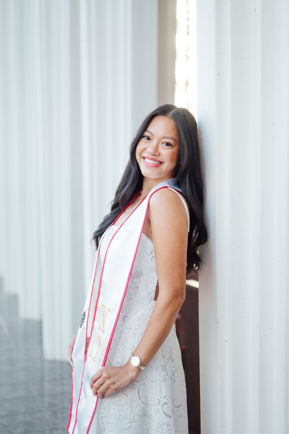 Smiling young woman in a white lace dress and graduation stole leaning against a white pillar.