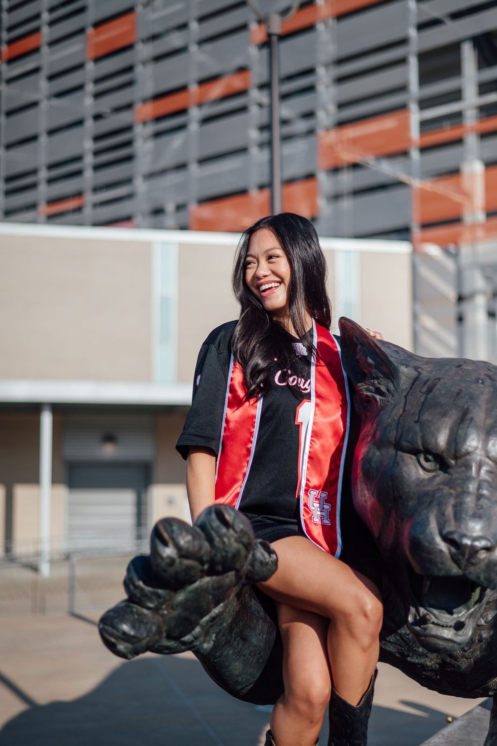 Smiling young woman in graduation sash sits on a panther statue outside a modern building.