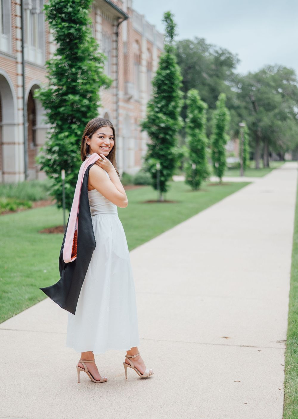 A smiling young woman in a light blue midi dress holds a graduation cap and stole on a tree-lined campus walkway.