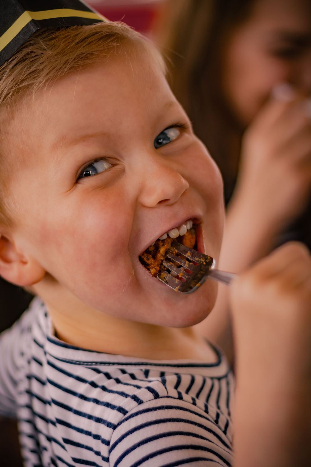 A young person in a striped shirt takes a big bite of chocolate cake with joy and enthusiasm.
