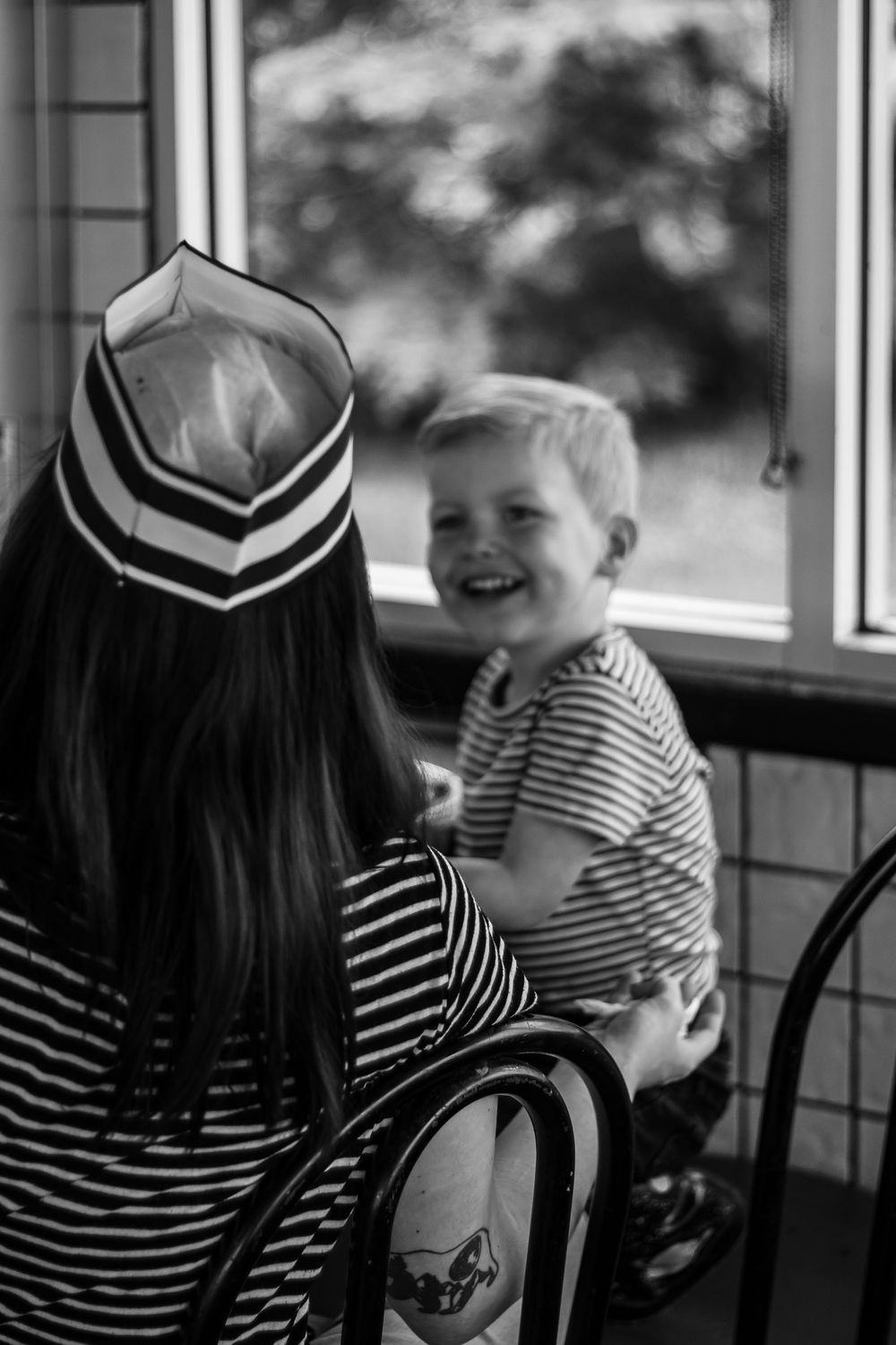 A black and white photo of a child smiling in a diner booth while someone in a striped shirt and cap sits nearby.