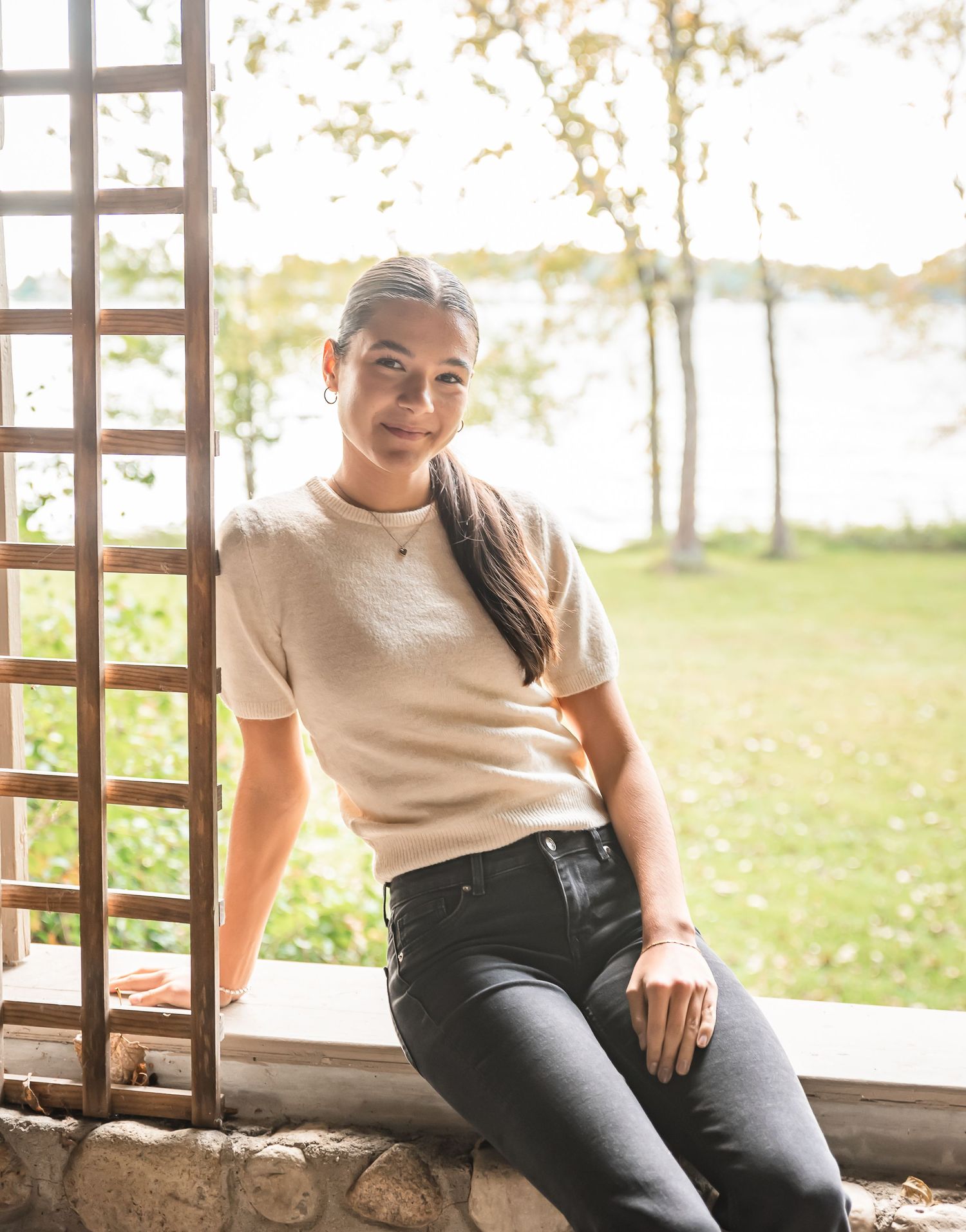 A casual portrait of someone sitting on a wooden ledge outdoors wearing a cream sweater and dark jeans.