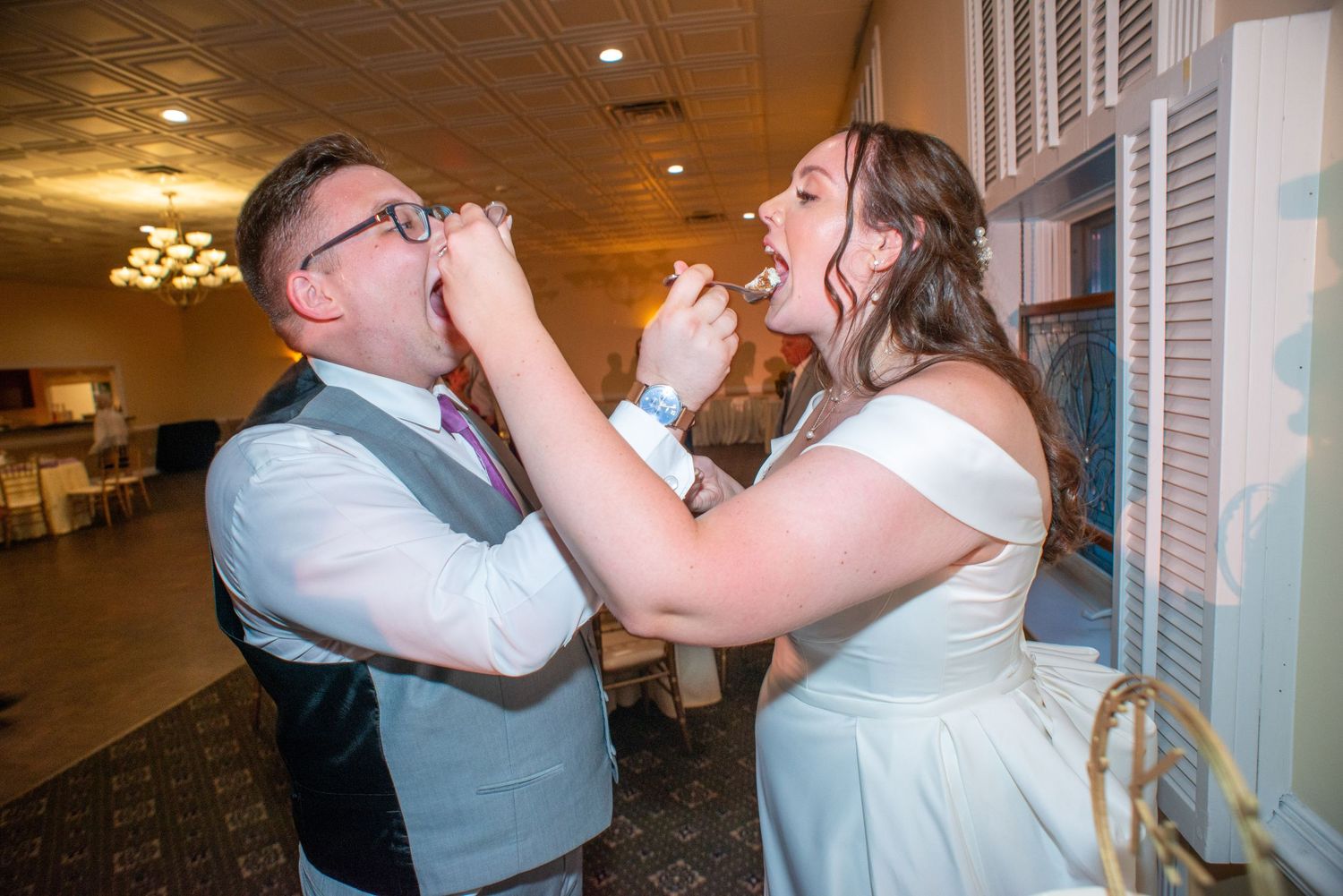 A couple feeds each other cake at their wedding reception in an indoor venue.