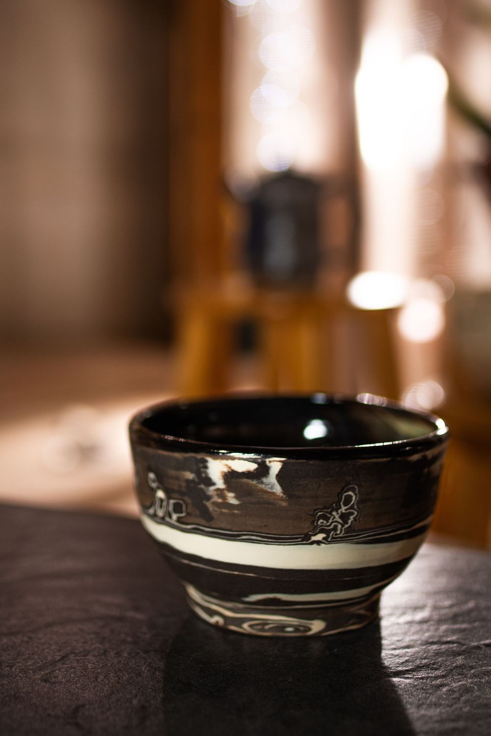 Dark glazed ceramic bowl with white swirl patterns on slate surface with bokeh background.